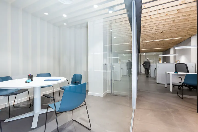 Sala de reuniones de las oficinas de Cuairón en Zaragoza, con mesa redonda blanca y sillas azules tras una mampara de vidrio