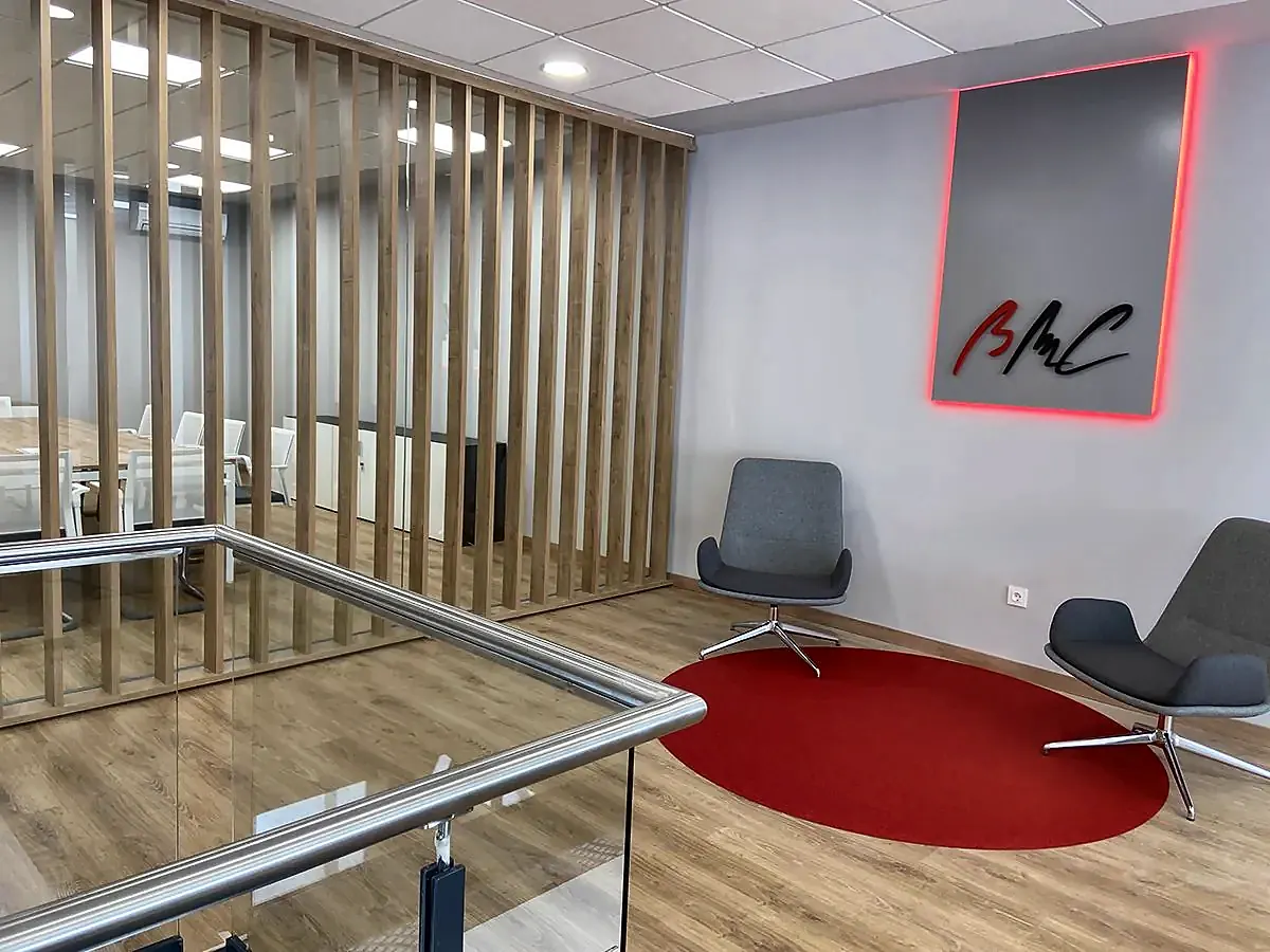 Rellano con butacas grises sobre alfombra roja, celosía de madera y logotipo retroiluminado