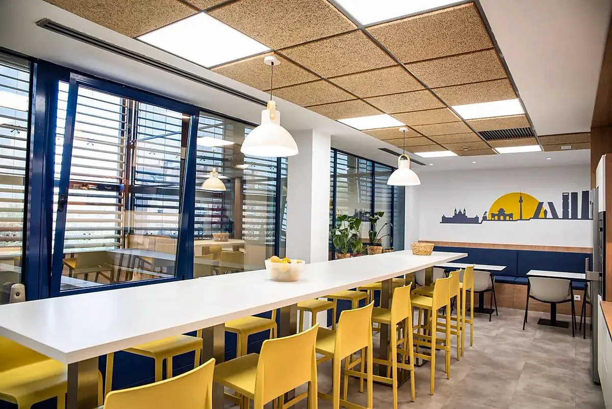 Office de la sede de Chint en Madrid: barra blanca con taburetes amarillos, ventanales con lamas y techo acústico