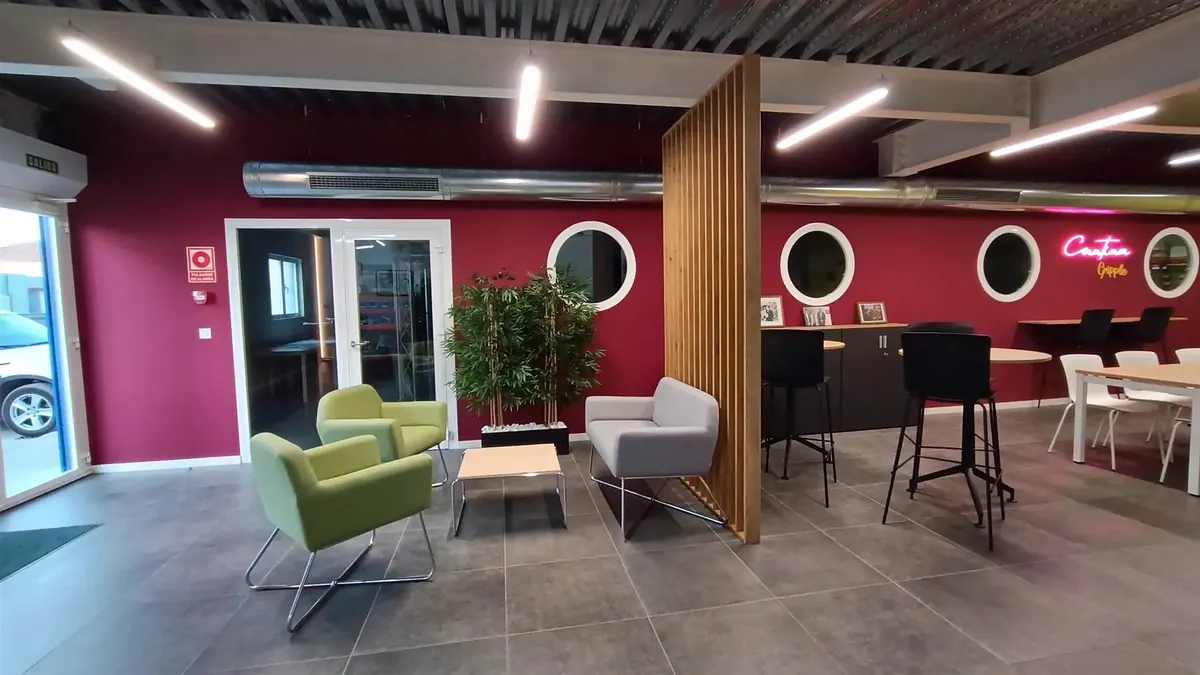 Zona social de Gripple en Zaragoza: butacas verdes y grises, celosía de madera y pared granate con ventanas de ojo de buey