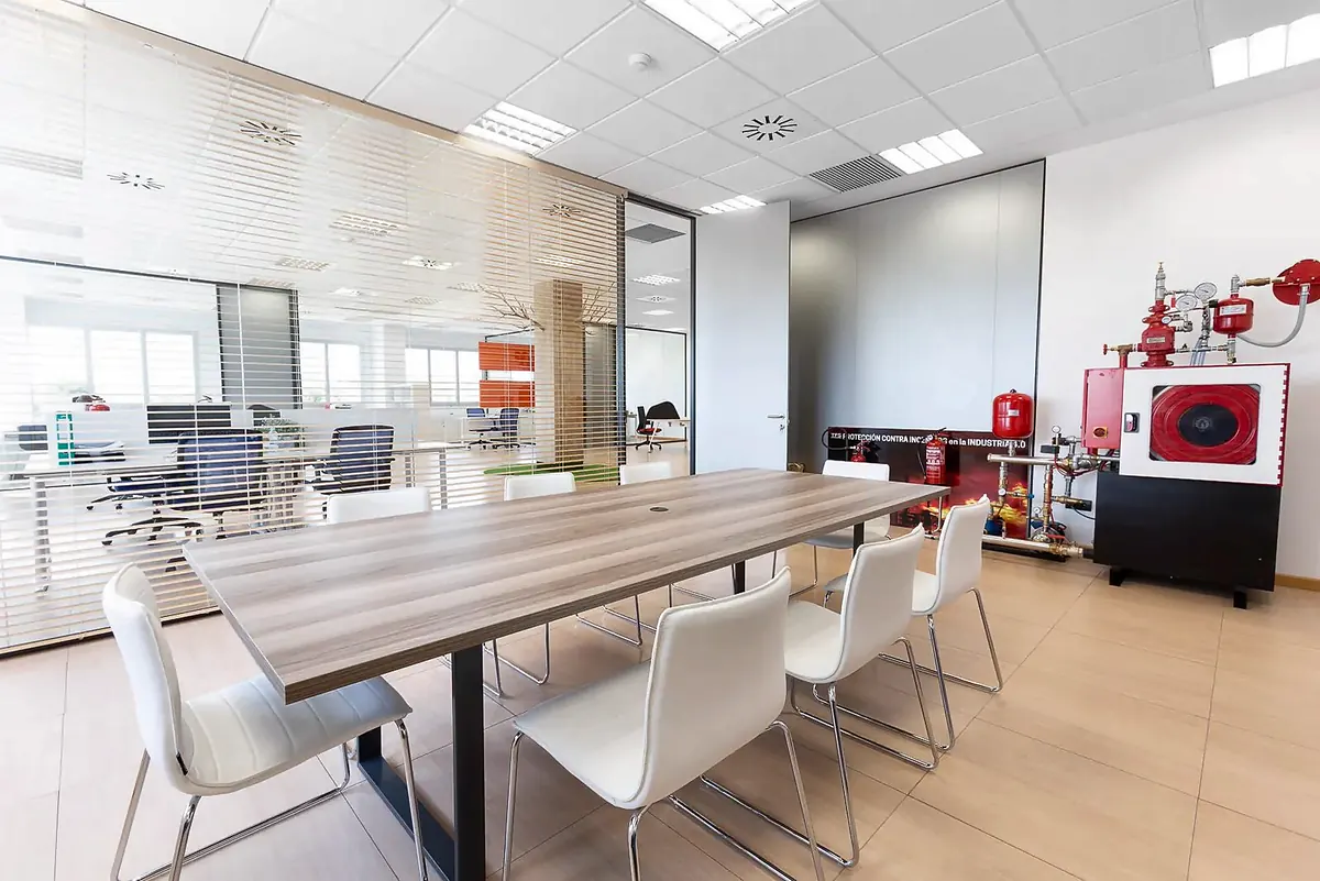 Sala de juntas con mesa de madera y sillas blancas junto a los despachos acristalados
